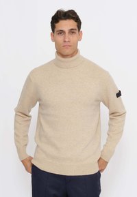 Maglione a collo alto beige realizzato in morbido tessuto a maglia. Presenta polsini e orlo a coste, con una piccola etichetta nera sulla manica sinistra.