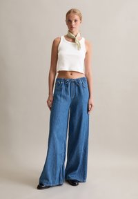 Un crop top blanc à côtes associé à un pantalon en denim bleu ample avec une taille élastique et un cordon de serrage. Le mannequin porte également une écharpe vert clair.