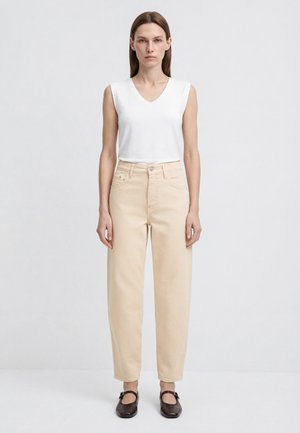 Frau mit glatten braunen Haaren, die ein weißes ärmelloses V-Ausschnitt-Oberteil, beige hoch taillierte Hosen und dunkelbraune Mary Jane Schuhe trägt und stillsteht.