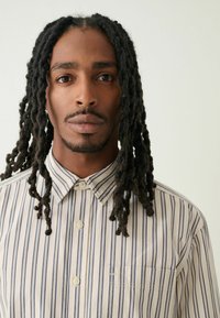 Košile s knoflíky s vertikálními modro-bílými pruhy, vyrobená z měkké látky, s límečkem a náprsní kapsou. Model má dlouhé, tmavé, texturované vlasy.