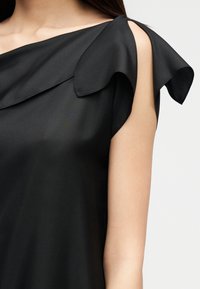 Femme portant un haut noir sans manches avec un décolleté asymétrique à volants et un détail sur l'épaule sur un fond clair.