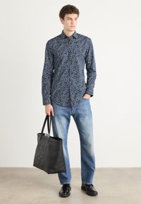 Blue patterned shirt with white floral design, paired with light blue jeans. Black tote bag with embossed logo and black shoes.