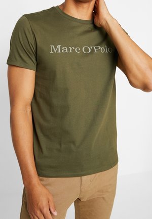 Mann trägt ein olivgrünes Marc O'Polo T-Shirt und beige Hose, mit einer Hand, die seinen Hals berührt, und der anderen Hand an seiner Seite.