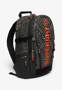 Czarny plecak z szarymi wzorzystymi sekcjami z napisem "SUPERDRY". Posiada pomarańczowe uchwyty zamków i liczne kieszenie na zamek.