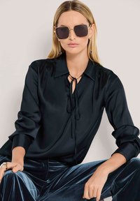 Schwarze Satinbluse mit Kragen und Schleifendetail, kombiniert mit weichen blauen Samthosen. Model trägt übergroße Sonnenbrille mit goldfarbenem Rahmen.