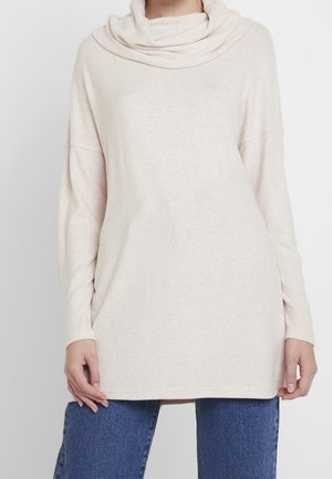 Femme portant un pull à col roulé ample, à manches longues, blanc cassé, associé à un jean bleu sur un fond clair uni.