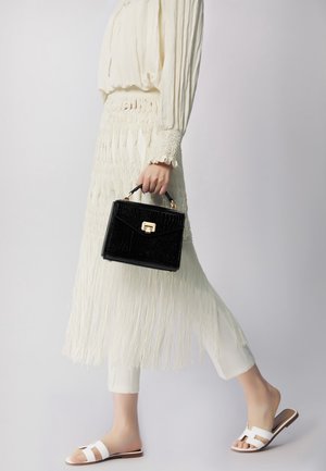 Sac à main en simili croco noir avec des accessoires dorés, associé à une jupe blanche à franges et des sandales blanches à glisser.