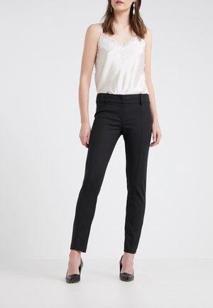 Femme portant un caraco en satin blanc orné de dentelle, un pantalon noir ajusté et des talons noirs, debout devant un fond uni.