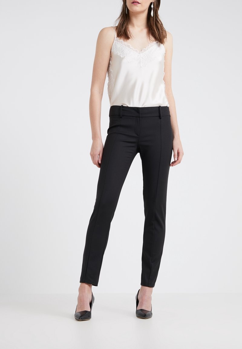 Femme portant un caraco en satin blanc orné de dentelle, un pantalon noir ajusté et des talons noirs, debout devant un fond uni.