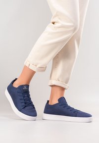 Granatowe siatkowe sneakersy z białą gumową podeszwą, z okrągłym noskiem, sześcioma otworami na sznurowadła i logo na języku. Teksturalny design cholewki.