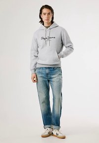 Sweatshirt cinzenta com capuz e bolso frontal, com o logótipo "Pepe Jeans", combinada com jeans azuis claros e ténis brancos com detalhes pretos.