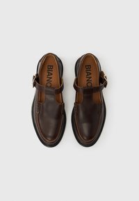 Ζευγάρι σκούρα καφέ δερμάτινων παπουτσιών Mary Jane με λουράκια με αγκράφες σε λευκό φόντο, με επενδύσεις ποδιών με την επωνυμία "BIANCO", θέα από πάνω.