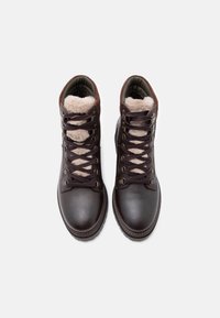 Barna bőr túrabakancs, puha szőrme béléssel, kerek orral és fémbetétekkel, fűzős kialakítással és texturált felülettel.