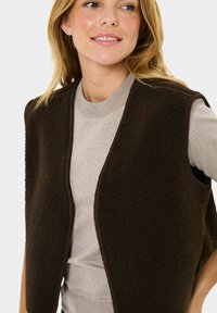 Joven mujer vistiendo una camiseta de manga larga beige y un chaleco de forro polar sin mangas marrón oscuro, sonriendo y mirando ligeramente de lado.