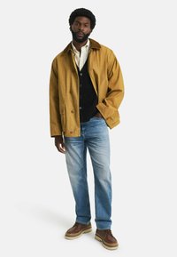 Hellbrauner Baumwolljacke mit Knopfverschluss, über einer schwarzen Weste und einem weißen Hemd getragen, kombiniert mit ausgebleichten blauen Jeans und braunen Schnürschuhen.