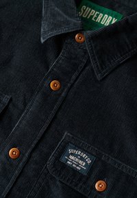 Ciemnoniebieska koszula z korduroy z zapięciem na guziki, dwoma kieszeniami na piersi oraz metką z napisem "SUPERDRY CO." w białym tekście na niebieskim tle.
