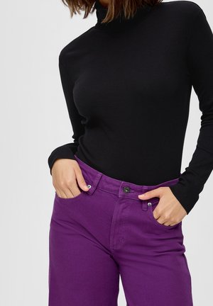 Vrouw draagt een zwarte coltrui met lange mouwen, ingestopt in een paar paarse high-waisted jeans, met handen deels in de zakken.