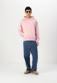 Sweat-shirt à capuche rose clair avec une poche avant, associé à un jean en denim bleu lâche et des chaussures beiges. Design décontracté, coupe relax.