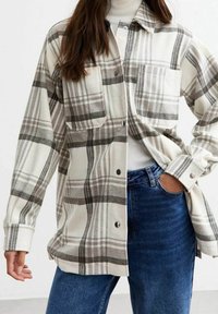 Camicia in flanella a quadri beige con motivi a griglia grigia e nera, dotata di due tasche frontali, chiusura a bottoni e vestibilità comoda.