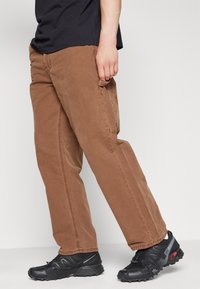 Pantalones cargo de algodón marrón con bolsillos laterales, patas rectas y costuras en contraste, combinados con zapatillas deportivas negras de suela texturada.