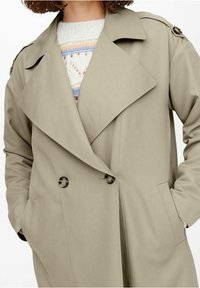 Femme portant un trench-coat beige croisé sur un pull en maille blanc à rayures colorées, les mains dans les poches du manteau.