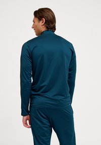 Långärmad träningsskjorta i mörk teal, med hög krage och slim fit. Slät yta med kontrasterande svarta sidodetaljer.