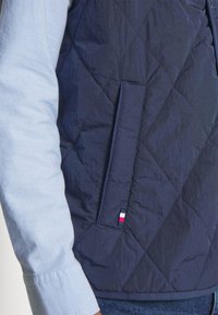 Marineblaues gesteppte Weste mit diagonaler Naht, ausgestattet mit einer Seitentasche und einem kleinen, markierten Akzent in Rot, Weiß und Blau am Saum.