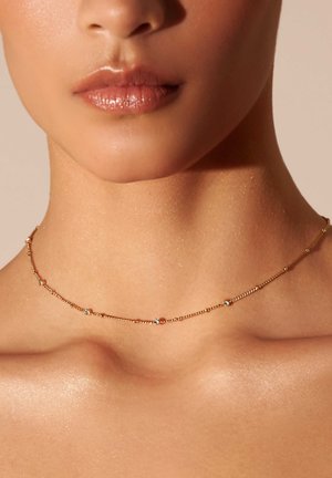 Collar de oro con pequeñas cuentas claras, que presenta un diseño de cadena delicado. El fondo es de un tono claro y sutil. La piel tiene un ligero brillo.