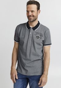 Sivé polo tričko s námorníckymi akcentmi, krátkymi rukávmi a golierom. Obsahuje biele lemovanie, potlač loga na prednej strane a červenú nášivku na ľavom rukáve.