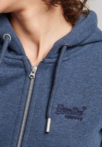 Sudadera con cremallera azul hecha de tela suave, con cremallera plateada, cordones y el logo bordado de "SuperDry" en púrpura oscuro.