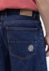 Tumši zili džinsu šorti ar oranžām stitching. Iespējas aizmugurējā kabata ar baltu ziedu un smaidīgu seju izšuvumu, kā arī brūns ādas tags.