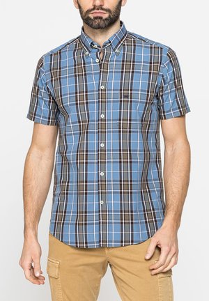 Uomo che indossa una camicia a quadri blu e marrone a maniche corte con pantaloni cargo kaki su uno sfondo uniforme.