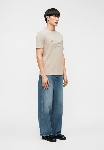 Νεαρός άνδρας όρθιος, φοράει μπεζ κοντομάνικο πουκάμισο με ανάγλυφο λογότυπο, μπλε τζιν με φαρδύ πόδι και μαύρα παπούτσια σε απλό φόντο.