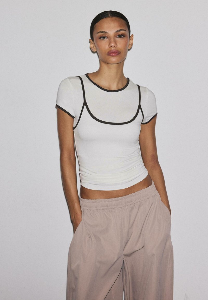 Jeune femme les mains dans les poches, portant un haut blanc ajusté à manches courtes avec un liseré noir et un pantalon beige ample, sur fond uni.
