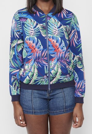 Nainen, joka pitää yllään sinistä kukallista vetoketjullista takkia, jossa on trooppisten lehtien kuviot, ja farkkushortseja, seisomassa yksiväristä taustaa vasten.