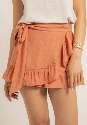 Jupe portefeuille en orange rouille, avec un tissu texturé, un ourlet volanté et un détail de ceinture nouée sur le côté.