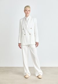 Blazer bianco doppiopetto con bottoni dorati, abbinato a pantaloni coordinati. Tessuto liscio e leggero. Scarpe casual con design testurizzato.