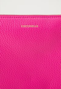 Fuchsia lærpung med struktureret overflade og et gyldent "Coccinelle" logo i midten, med lynlåslukning øverst.