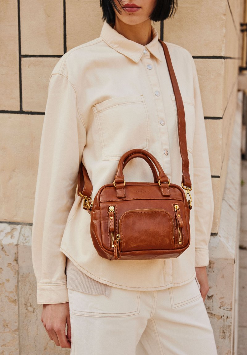 Sac à main en cuir marron avec deux poignées et une bandoulière amovible, doté de fermetures éclair dorées et d'une poche avant. Texture lisse, design compact.