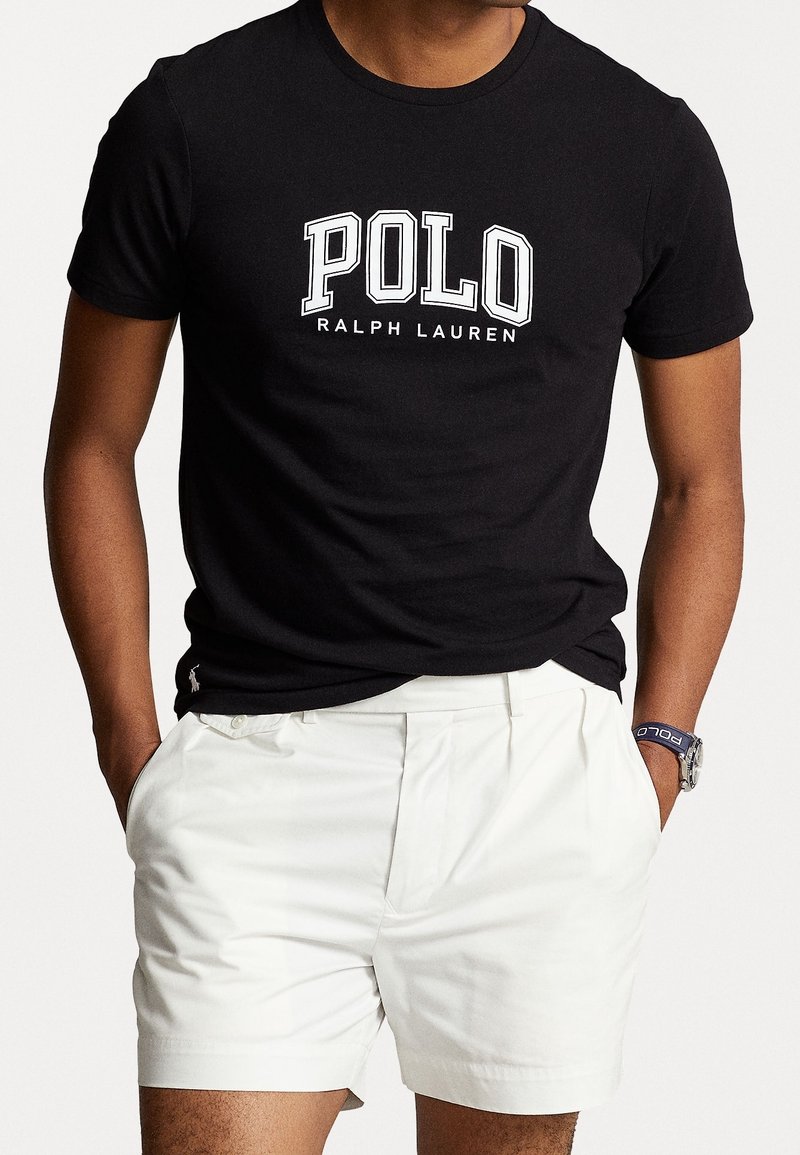 Muž oblečený v černém tričku Polo Ralph Lauren a bílých šortkách, ruce v kapsách, s hodinkami značky Polo.