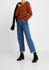 Pull marron avec un motif de teckel noir, associé à un jean droit bleu et des bottines noires. Comprend une veste noire.