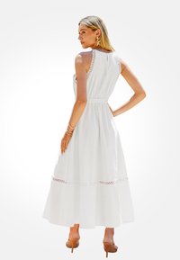 Biała maxi sukienka z dopasowaną talią, otwartym plecami i misternymi koronkowymi detalami wzdłuż ramion i dołu. Gładki materiał, długość do kostek.