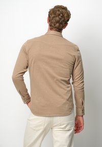 Camicia a maniche lunghe marrone chiaro con colletto, dettaglio della yoke posteriore e polsini con bottoni. Texture liscia e orlo arrotondato. Indossata con pantaloni bianchi.
