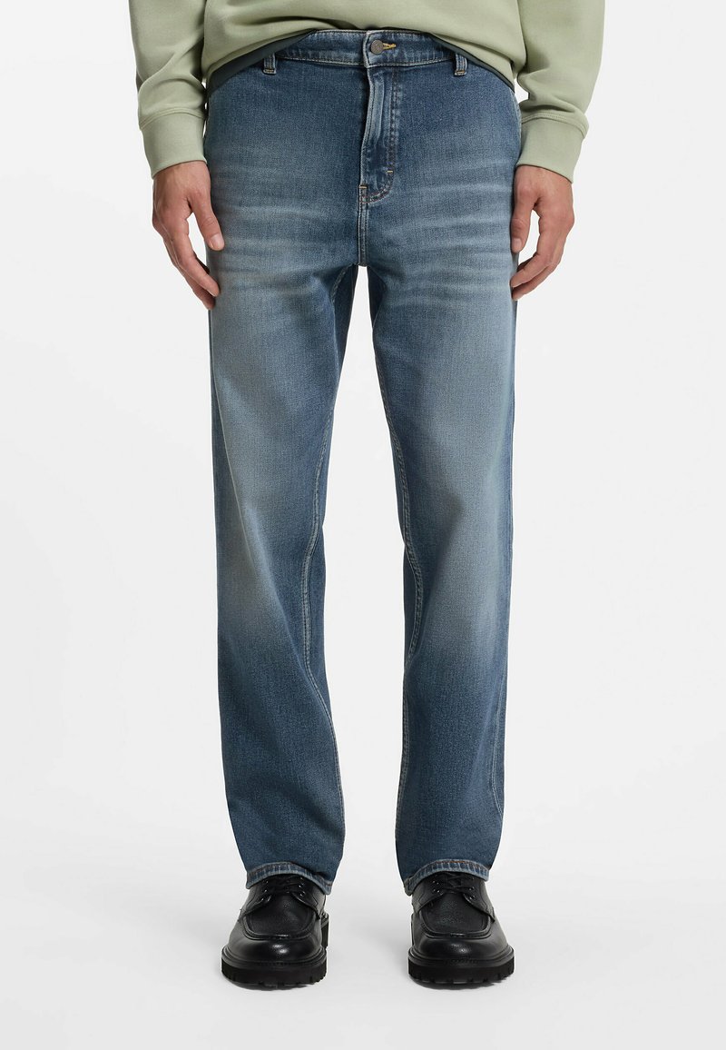 Homme portant un jean bleu clair délavé, des chaussures noires et un sweat-shirt vert, debout devant un fond blanc uni.