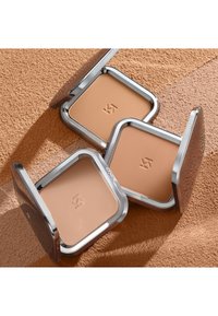 Drie open compactpoederdozen met beige tinten op een gestructureerd oppervlak, elk doosje gegraveerd met een minimalistisch logo.