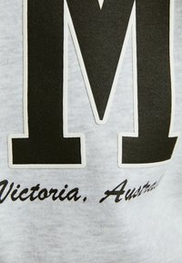 Felpa grigia con una grande lettera nera "M" e il testo "Victoria, Australia" in corsivo sotto, con un tessuto testurizzato.