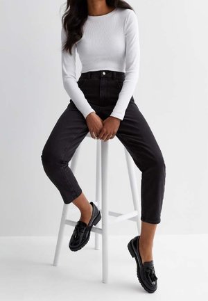 Woman wearing a white long-sleeve top, high-waisted black jeans, and black tassel loafers sitting on a white stool against a plain backdrop.
