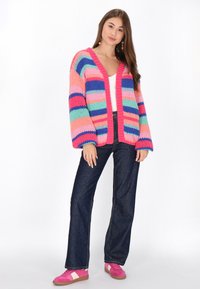 Strickjacke mit kräftigen horizontalen Streifen in Pink, Blau, Grün und Lila. V-Ausschnitt-Design mit weiten Ärmeln, getragen über einem weißen Oberteil.
