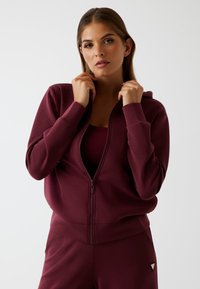 Vrouw in een bordeauxrode zip-up hoodie en bijpassende broek, houdt de kraag van de hoodie met beide handen vast tegen een effen achtergrond.
