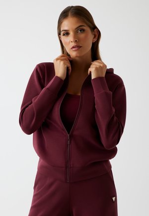 Vrouw in een bordeauxrode zip-up hoodie en bijpassende broek, houdt de kraag van de hoodie met beide handen vast tegen een effen achtergrond.